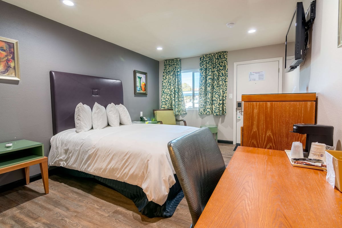 A hotel room with a large bed, a wooden desk, a television mounted on the wall, and a window with curtains.