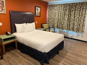 A hotel room with a large bed, a bedside table with a phone, a chair, a desk, and a window with curtains.