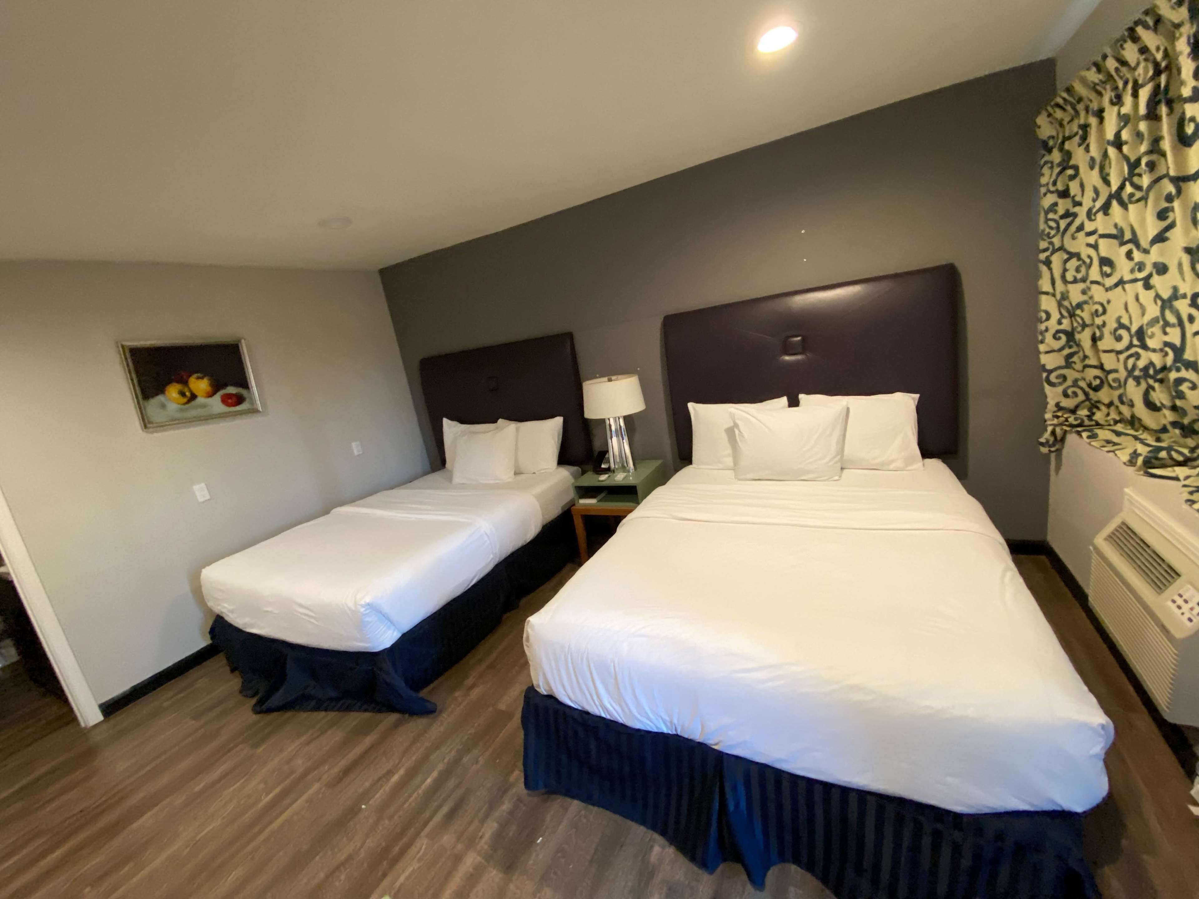 A hotel room with two single beds, a nightstand with a lamp, a small painting on the wall, and a window with patterned curtains.