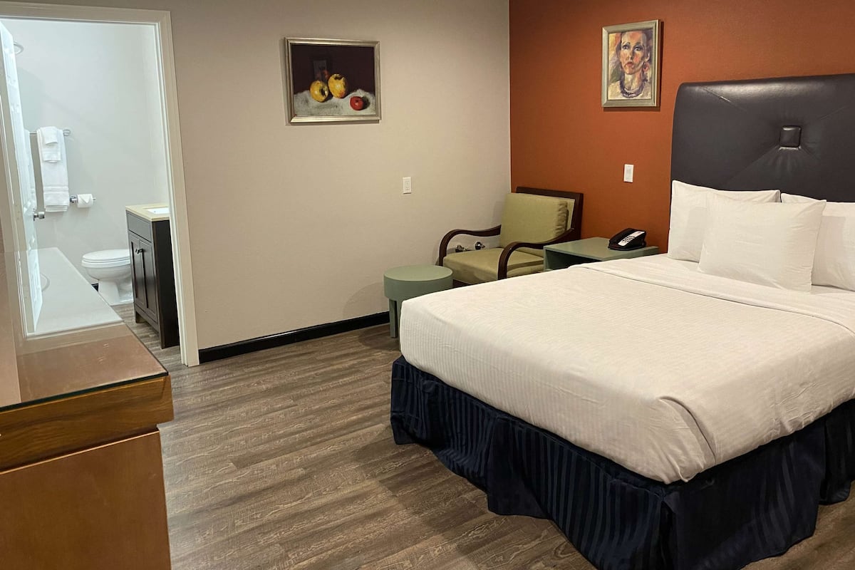 A hotel room with a bed, a chair, a small table, a phone, and a painting on the wall.
