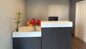 A reception desk with a white top and dark side panel, a potted bamboo plant, and a vase with colorful flowers. - Motel 6 Ridgecrest, CA - China Lake (Ridgecrest)