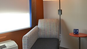 Ein kleines Zimmer mit einem Einzelbett, einer karierten Bettdecke, einer Stehlampe, einem kleinen Tisch und einem Fenster mit Jalousien.