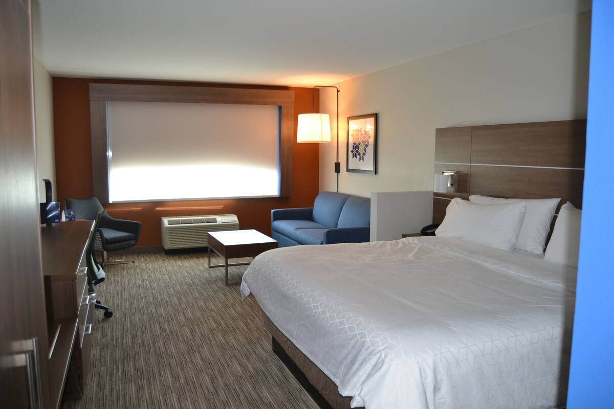 A hotel room with a large bed, a desk, a chair, a sofa, a TV, and a window with blinds.