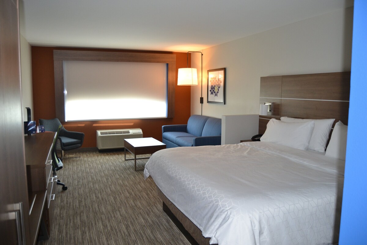 A hotel room with a large bed, a desk, a chair, a small table, a lamp, and a window with blinds.