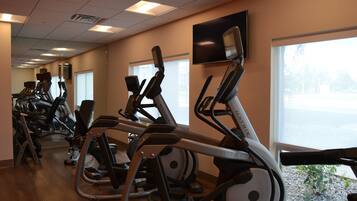 A row of stationary exercise bikes in a gym with large windows and a flat-screen TV on the wall.