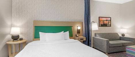 A hotel room with a bed, a chair, a nightstand, a lamp, and a painting on the wall.