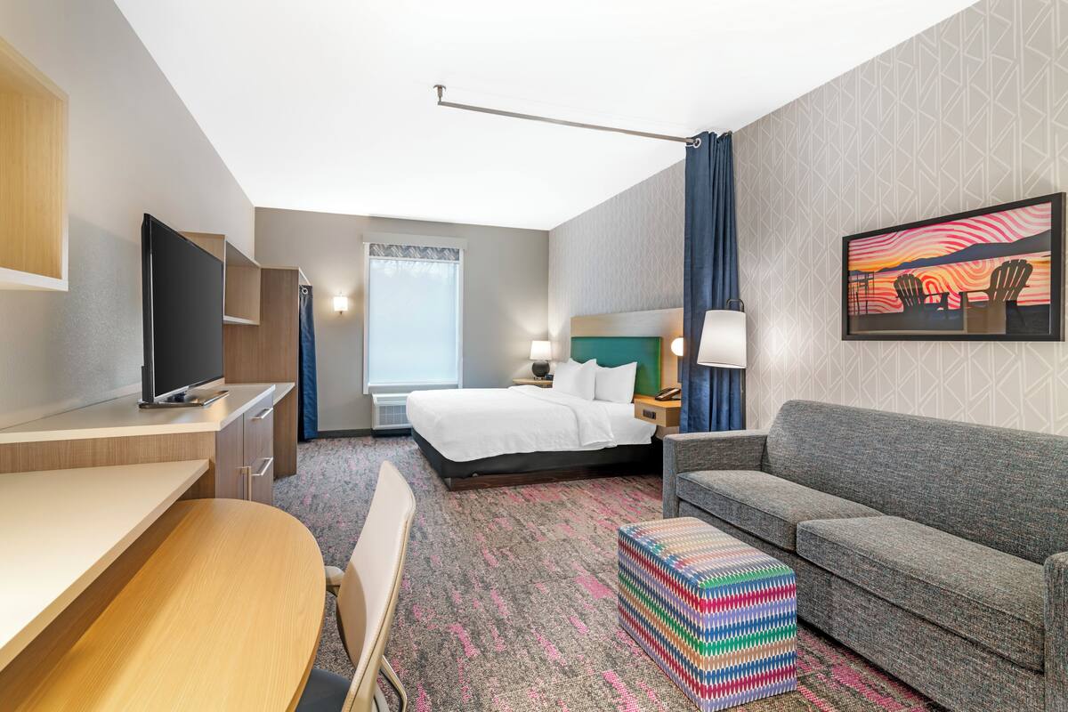 Een hotelkamer met een bed, een bureau, een slaapbank, een televisie, een raam met gordijnen en een schilderij aan de muur.
