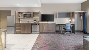 A modern kitchen with a refrigerator, microwave, and dishwasher, a flat-screen TV, a desk with a chair, and a patterned carpet. - Home2 Suites by Hilton Queensbury Lake George (Queensbury)