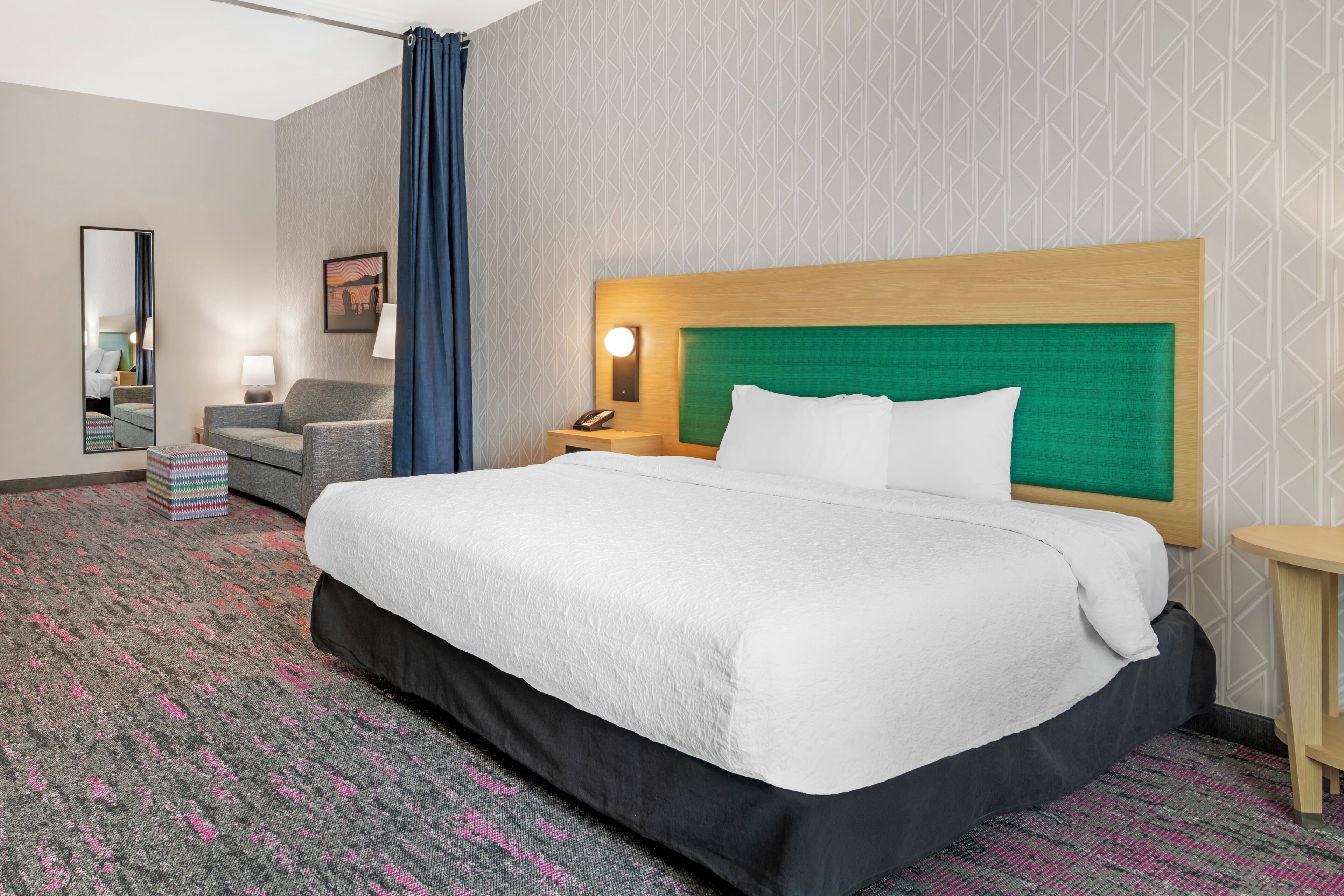 A hotel room with a large bed, a nightstand with a lamp, a small table, a chair, and a mirror.