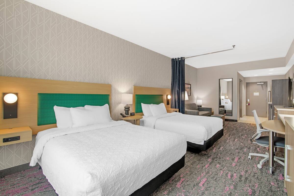 Een hotelkamer met twee bedden, een bureau, een stoel, een spiegel en een deur die naar een andere kamer leidt.