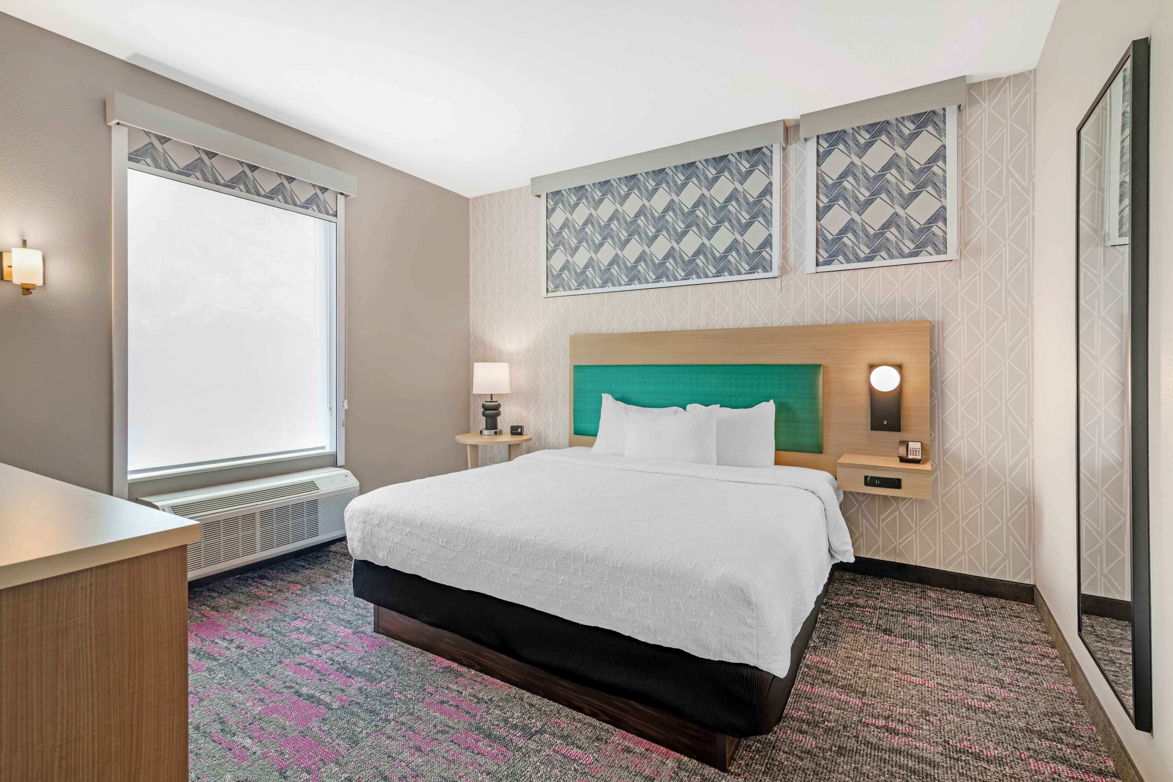 A hotel room with a bed, bedside tables, a desk, a window with blinds, and patterned wall decor.