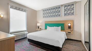 A hotel room with a bed, bedside tables, a desk, a window with blinds, and patterned wall decor.