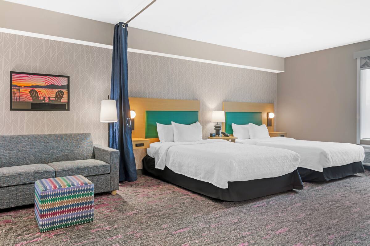 Een hotelkamer met twee bedden, een bank, een kleurrijk voetenbankje, een schilderij aan de muur en een groot raam.