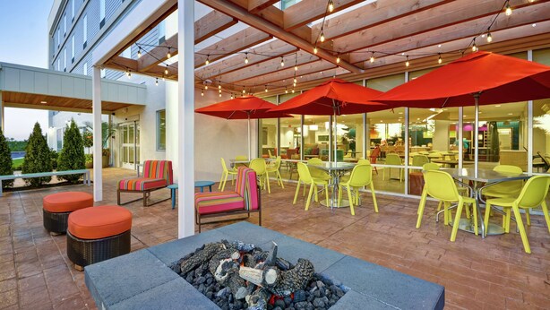 An outdoor patio area with a fire pit, yellow and red outdoor seating, and a covered wooden structure with string lights. - Home2 Suites by Hilton Queensbury Lake George (Queensbury)