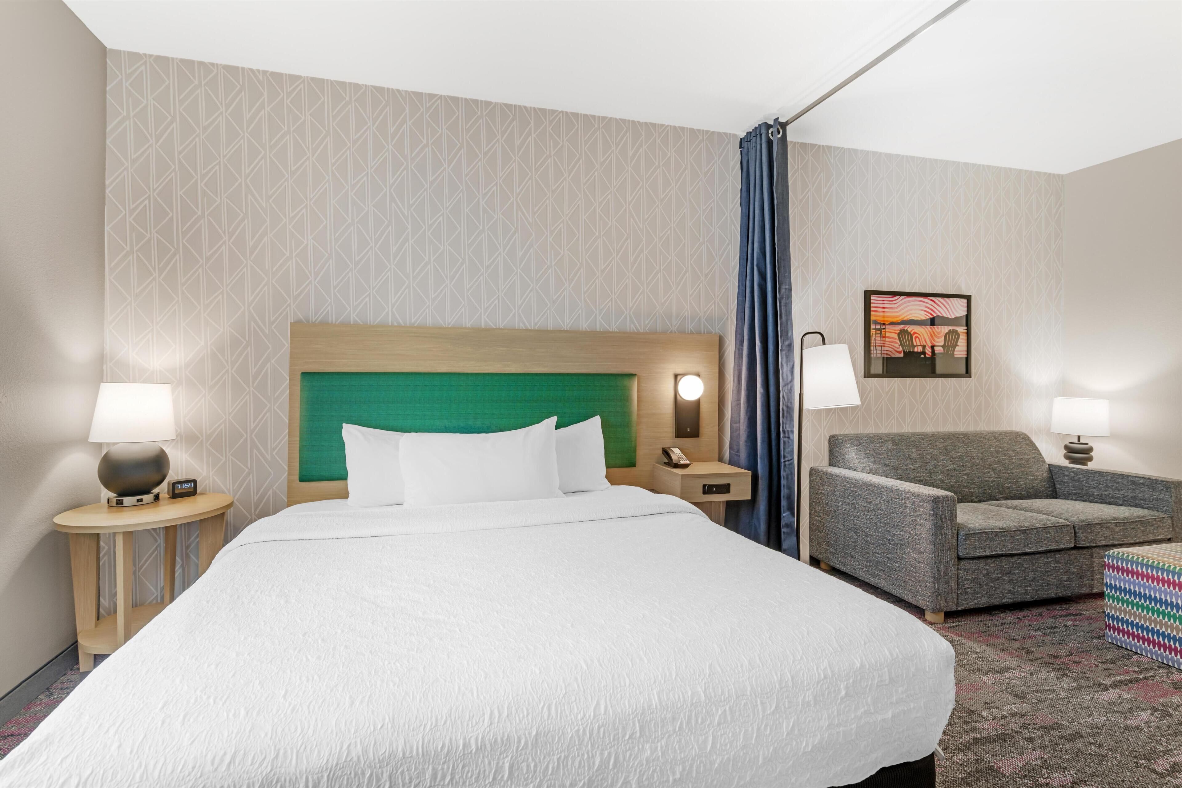 A hotel room with a large bed, a green headboard, two bedside tables with lamps, a gray armchair, and a framed picture on the wall.