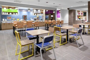 A modern cafeteria with wooden tables and chairs, a counter with food items, and a 'GOOD MORNING' sign. - Home2 Suites by Hilton Orlando South Park (Orlando)