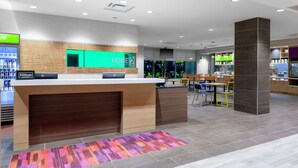 A hotel lobby with a reception desk, a green 'HOME2' sign, a minibar, and a seating area with chairs and tables. - Home2 Suites by Hilton Orlando South Park (Orlando)