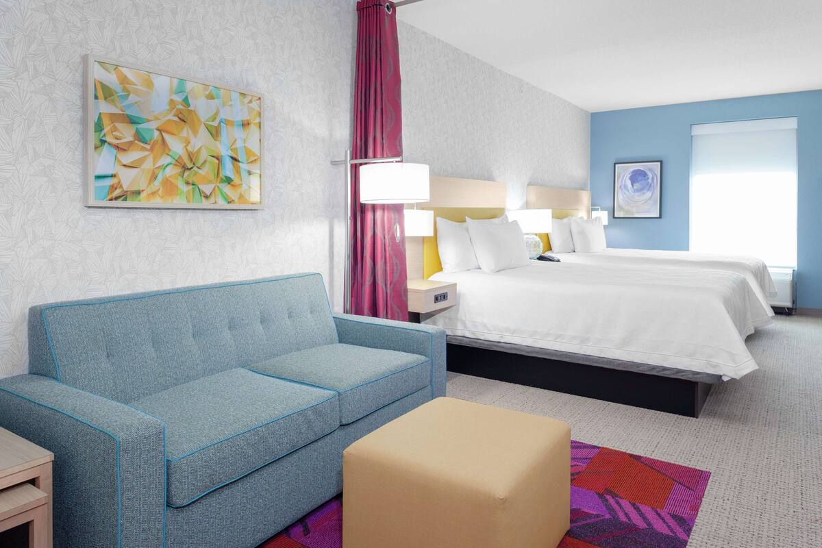 A hotel room with a blue sofa, a bed with white linens, a nightstand, a lamp, and a painting on the wall.