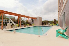 Una piscina con una pérgola de madera, sillas de descanso y un edificio al fondo.