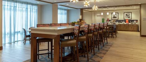 Uma área de refeições ampla, com uma mesa comprida e várias cadeiras, uma cozinha com bancada e eletrodomésticos, e uma grande janela com cortinas.