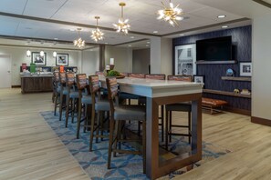 A modern hotel dining area with a long communal table, bar stools, and a television mounted on the wall. - Hampton Inn Bourbonnais Kankakee (Bourbonnais)