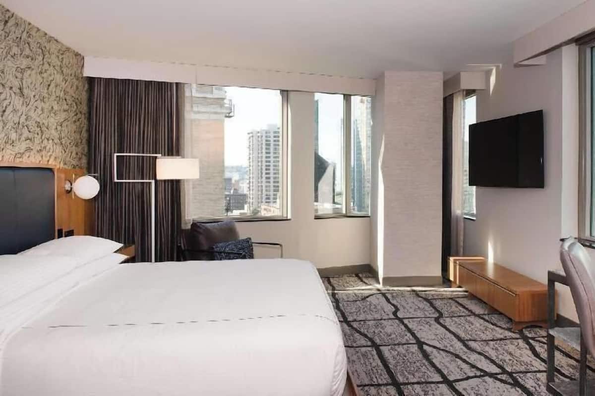 A hotel room with a bed, a desk, a chair, a TV, and large windows with a city view.