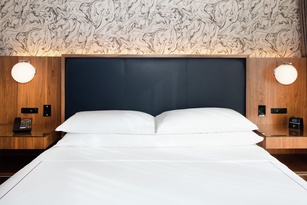 Une chambre d’hôtel avec un grand lit, une tête de lit en bois, deux tables de chevet avec des lampes et un papier peint à motifs.