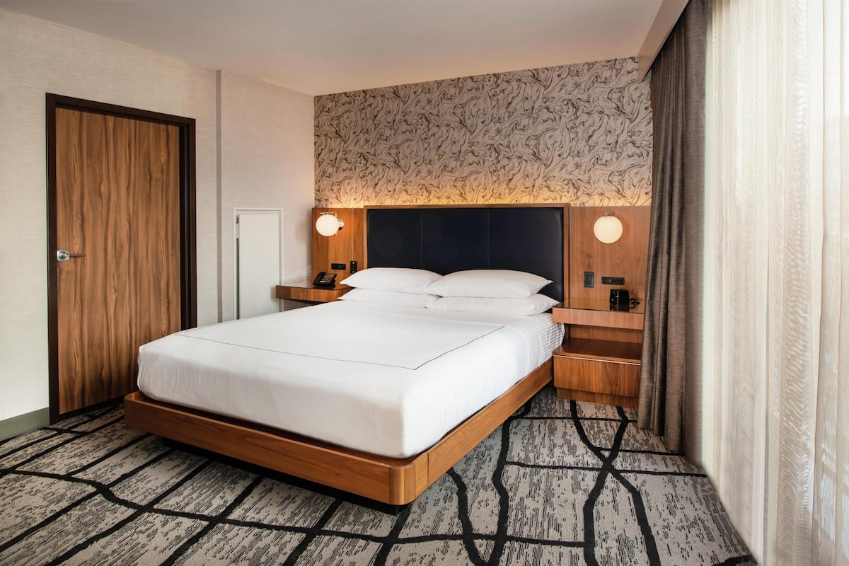 A hotel room with a large bed, wooden headboard, two bedside tables with lamps, a nightstand, a wall-mounted phone, and a patterned carpet.