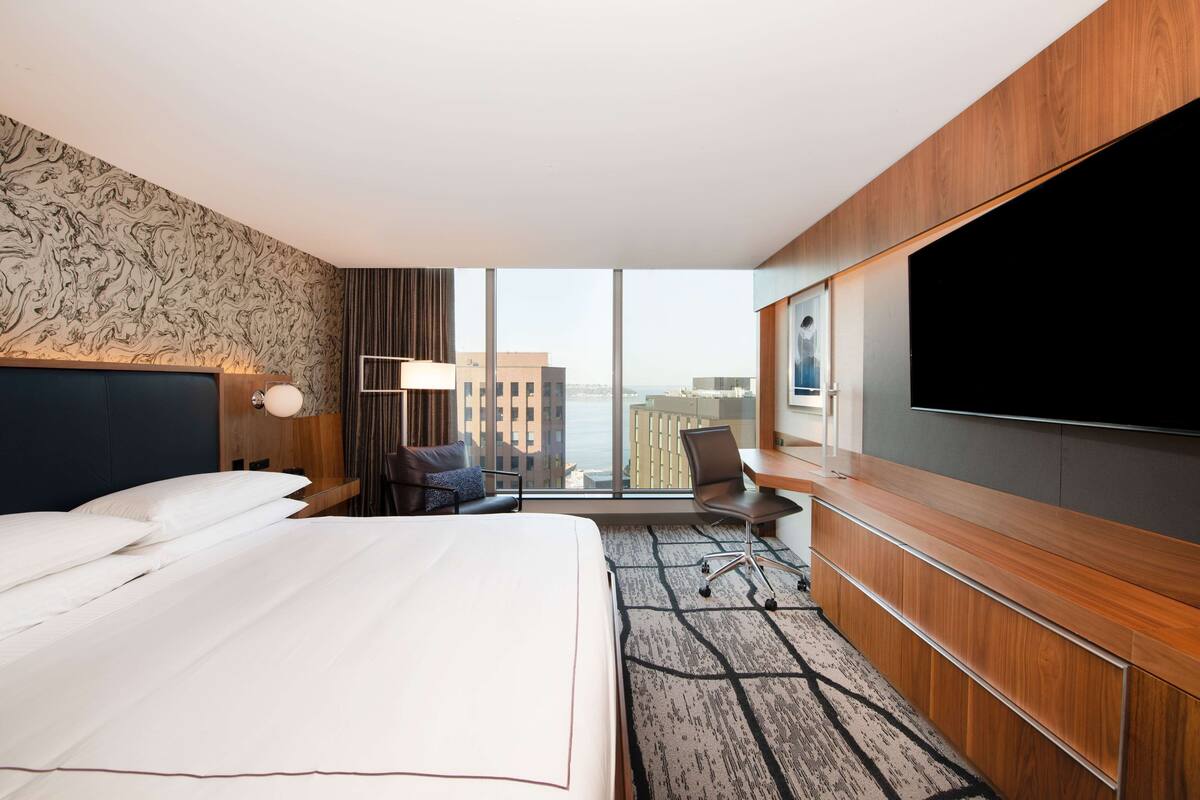 A hotel room with a large bed, a desk, a chair, a flat-screen TV, and a view of the city.
