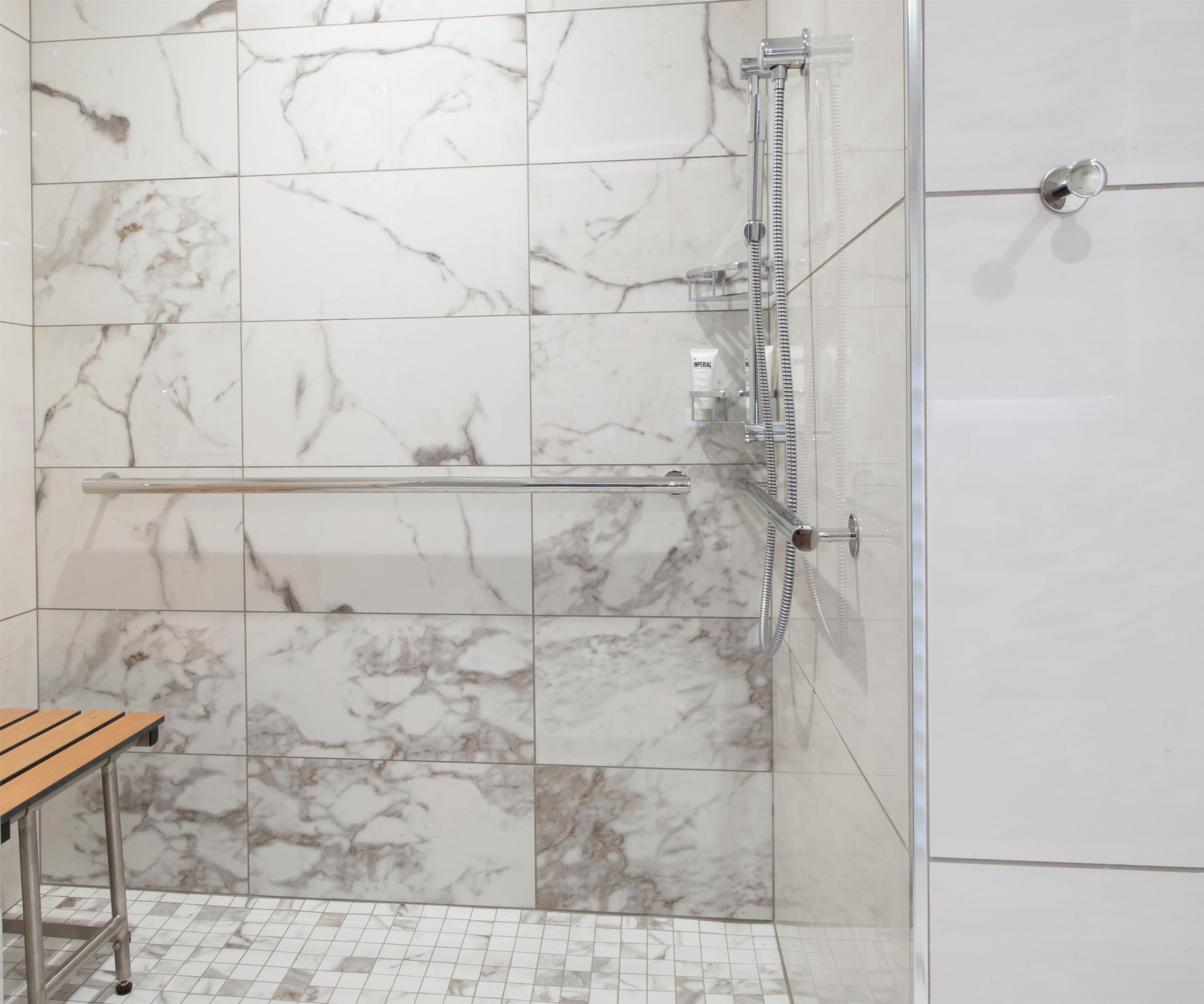 Un baño con baldosas de mármol, cabina de ducha de vidrio, toallero de metal y banco de madera.