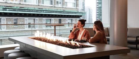 A modern bar area with a central fire feature, seating, and a view of the cityscape.