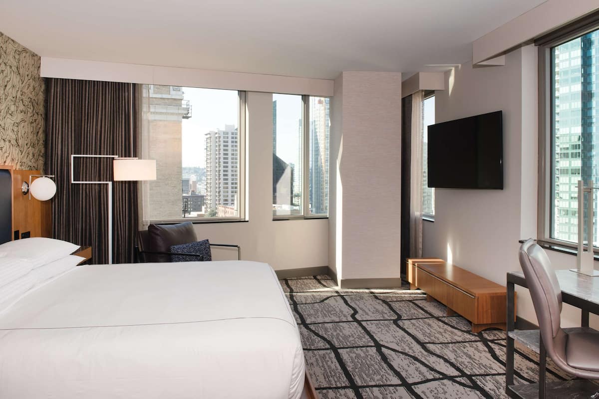 A hotel room with a large bed, a desk, a chair, a TV, and a view of the cityscape through large windows.