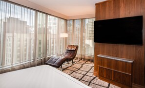 A hotel room with a large bed, a leather chair, a TV mounted on a wooden panel, and a view of buildings through sheer curtains. - The Charter Hotel Seattle, Curio Collection by Hilton (Seattle)