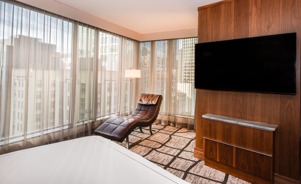 Une chambre d’hôtel avec un grand lit, un fauteuil en cuir, un téléviseur fixé sur un panneau en bois et une vue sur les immeubles à travers de fines rideaux.