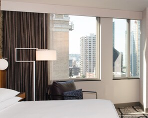 Une chambre d’hôtel avec un lit, une chaise, une lampe et une fenêtre donnant sur des immeubles imposants.