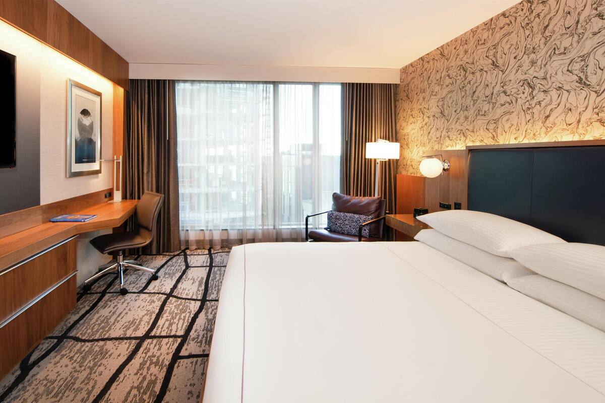 A hotel room with a bed, desk, chair, TV, and large windows.