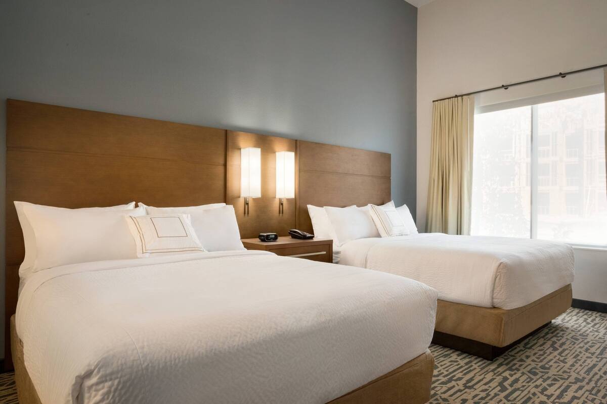 A hotel room with two beds, a wooden headboard, bedside lamps, a nightstand, and a large window with curtains.