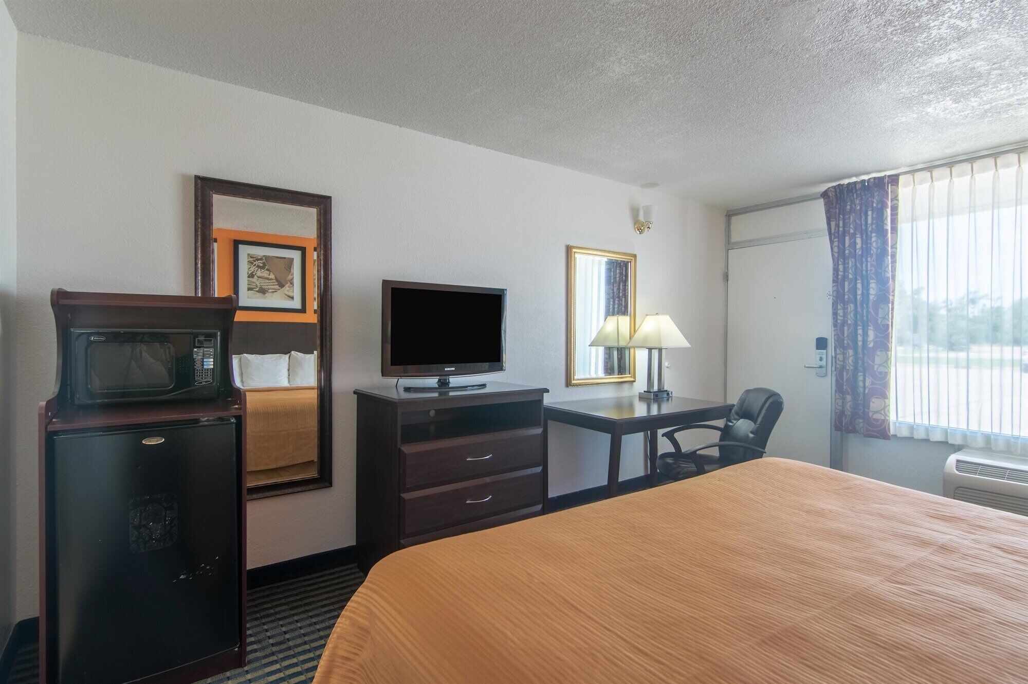 A hotel room with a bed, a desk, a television, a microwave, a chair, and a window with curtains.