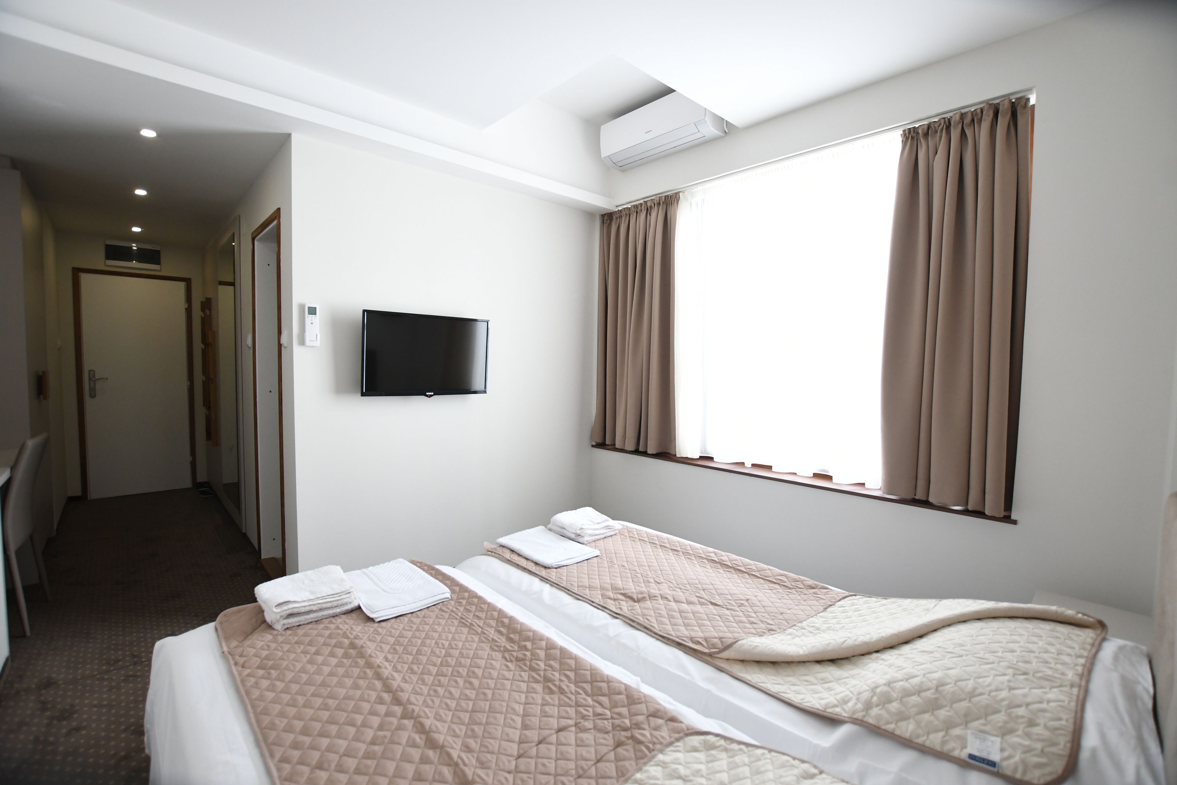 A hotel room with two beds, a flat-screen TV, an air conditioning unit, and a window with curtains.
