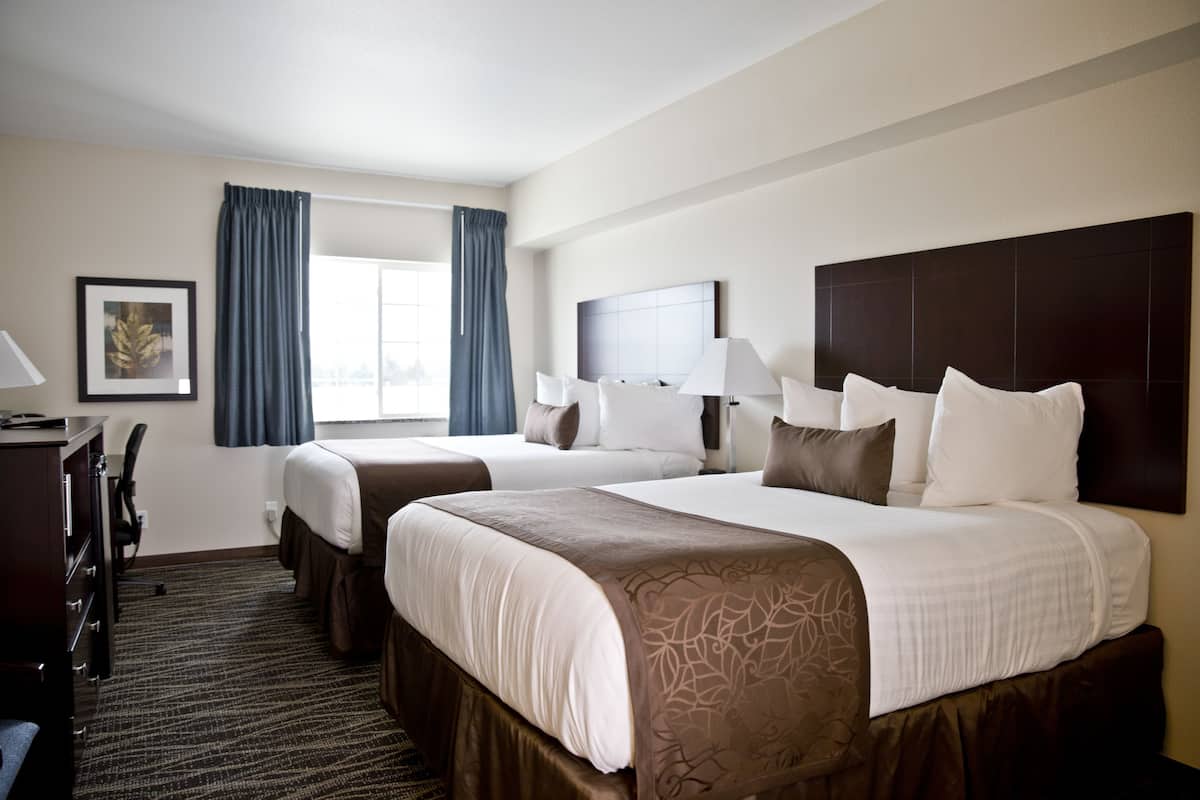 A hotel room with two beds, a desk, a chair, a window with curtains, and a framed picture on the wall.