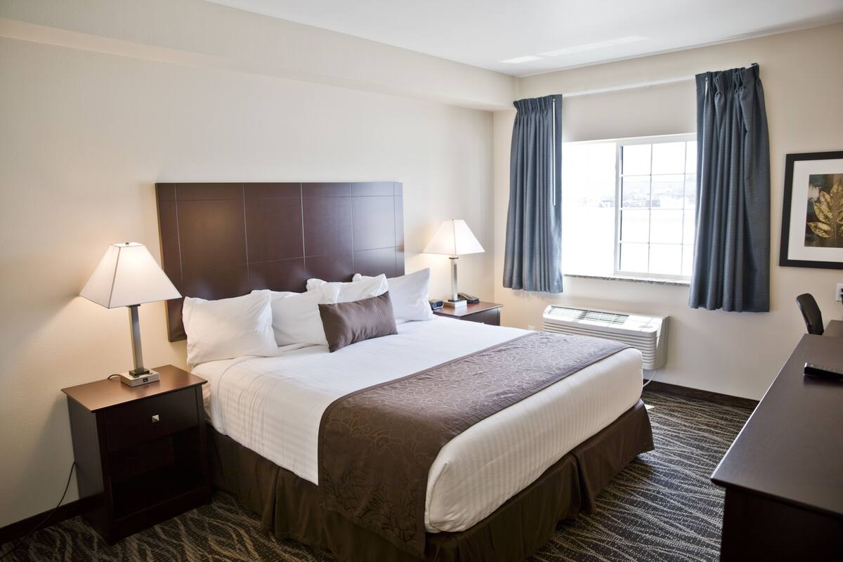 A hotel room with a large bed, two bedside tables with lamps, a desk, a window with curtains, and a framed picture on the wall.