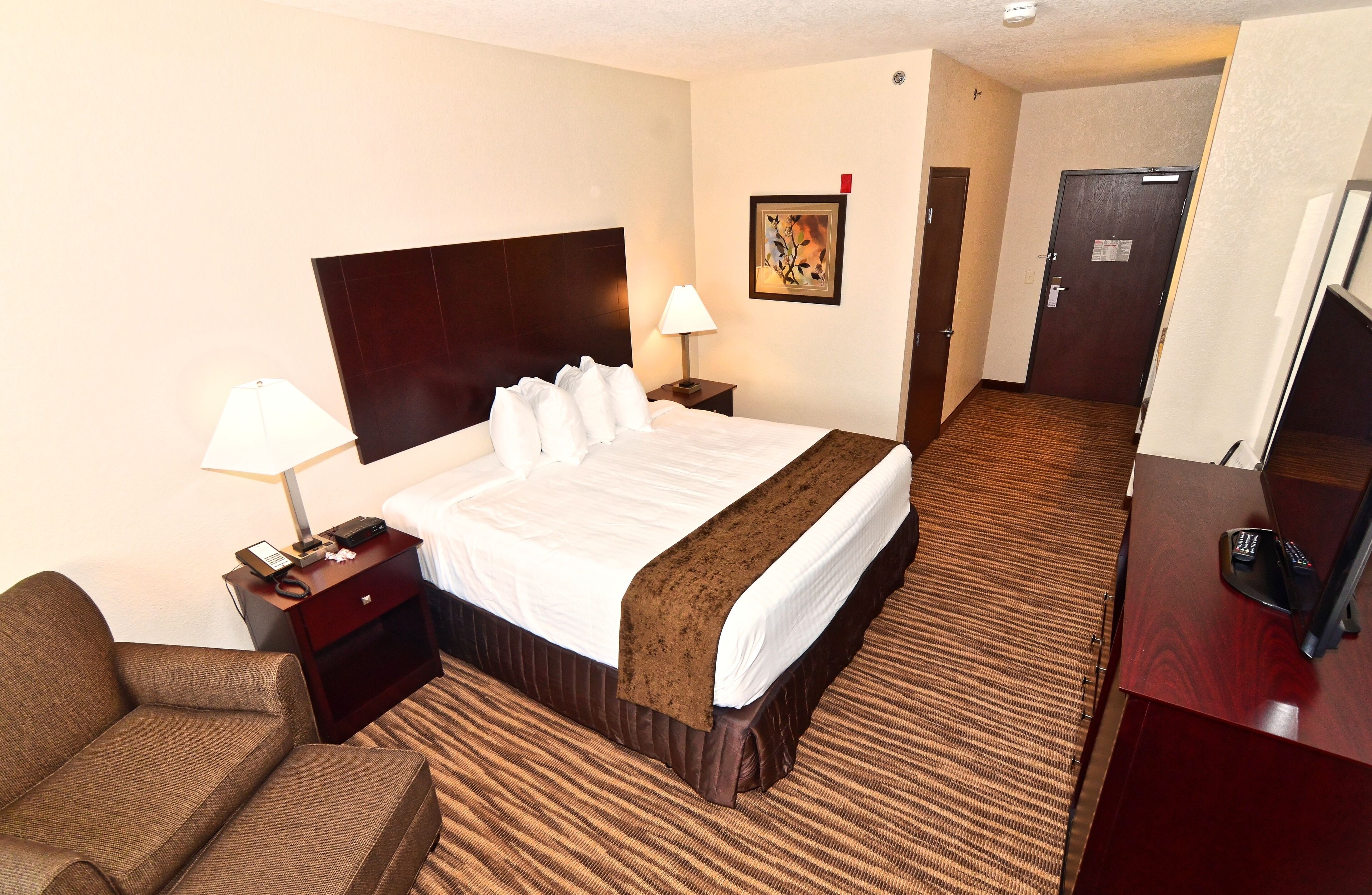 A hotel room with a large bed, a desk with a computer, a chair, a nightstand with a lamp, and a door.
