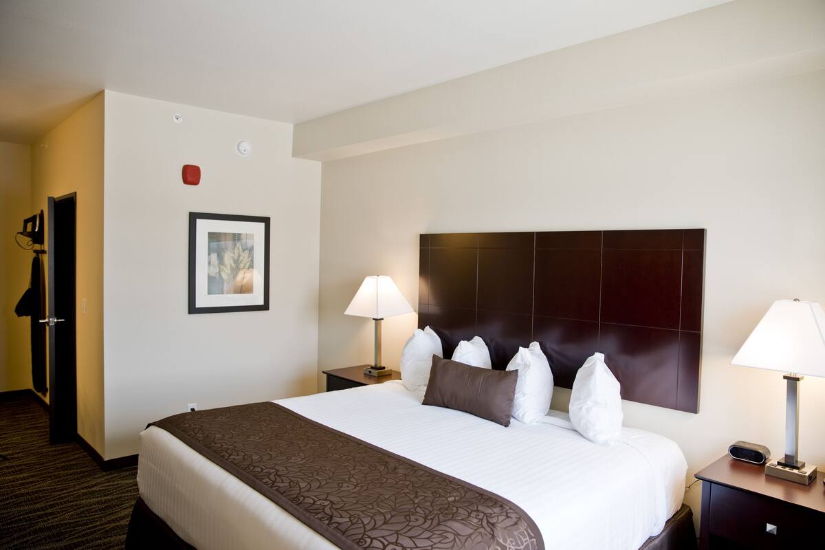 A hotel room with a large bed, two bedside tables with lamps, a headboard, a framed picture on the wall, and a coat rack.
