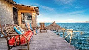 A wooden deck with wicker furniture, a thatched umbrella, and a view of the sea. - Uros LodgeTiticaca Peru (Puno)