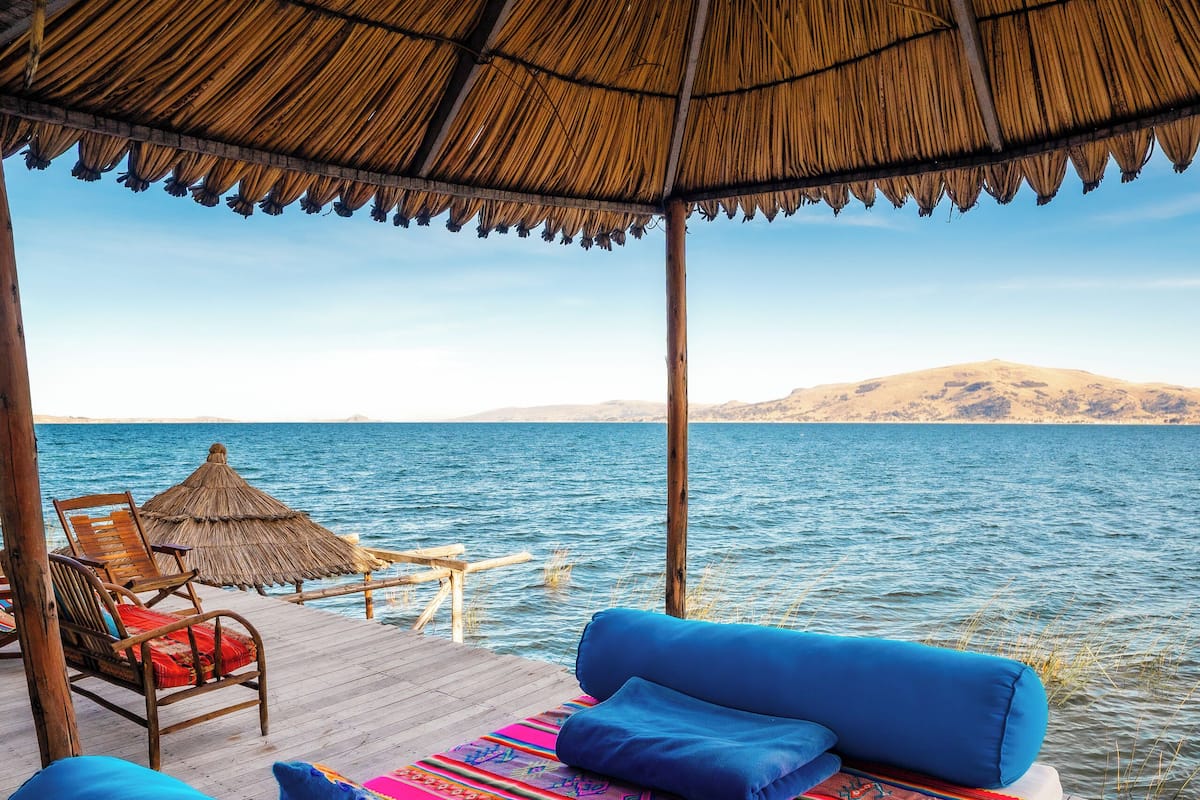 A wooden deck with a thatched roof, a bed with blue pillows, and a view of the sea.