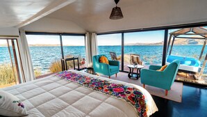 A bedroom with a large bed, a view of the sea, and two chairs by the window. - Uros LodgeTiticaca Peru (Puno)