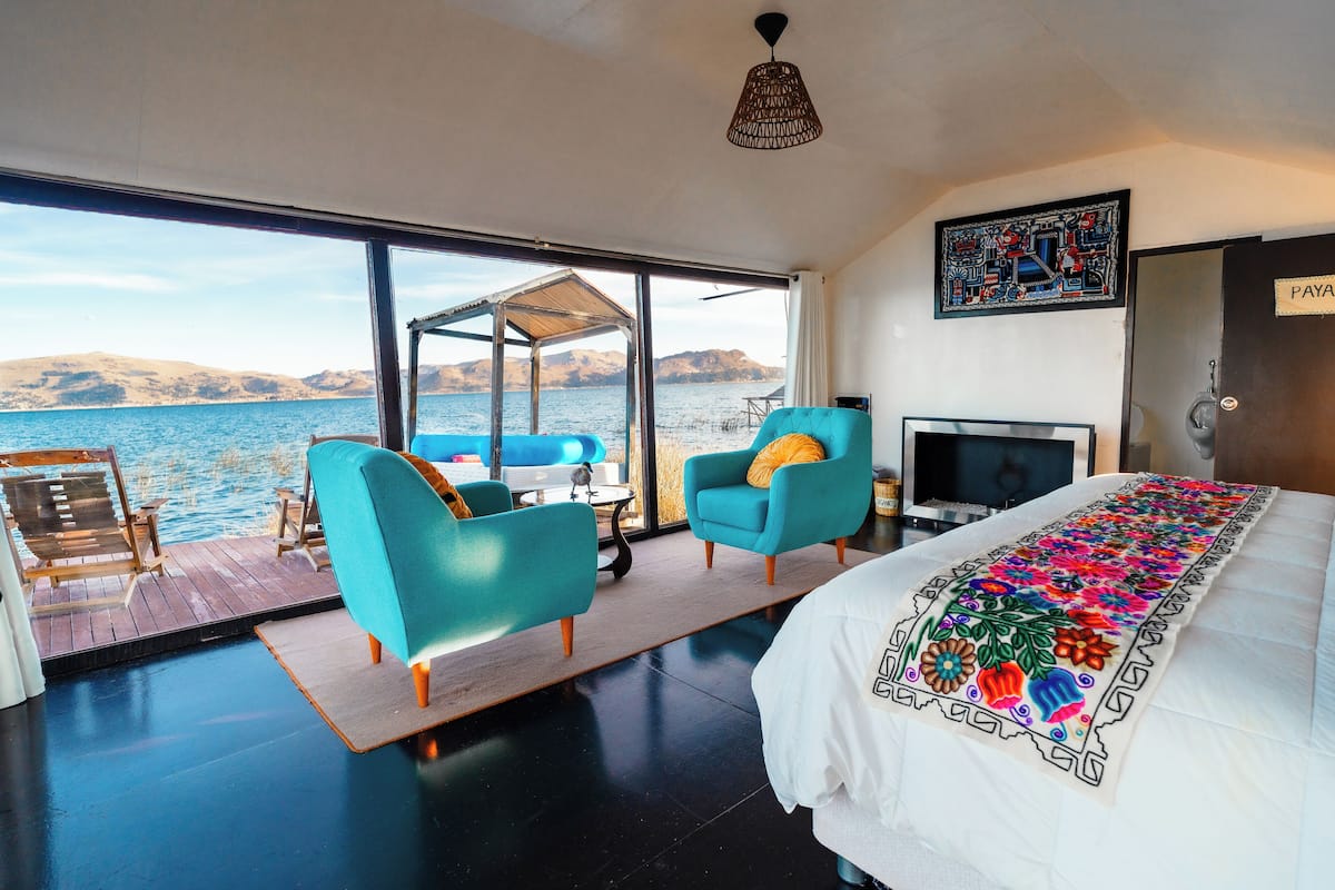 A room with a large window overlooking water, a bed with a colorful blanket, two blue armchairs, a small table, and a television.