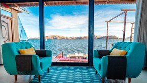 A room with two teal armchairs, a patterned rug, and a view of the sea and hills. - Uros LodgeTiticaca Peru (Puno)