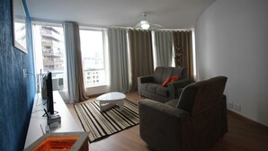 Ein modernes Wohnzimmer mit Sofa, Couchtisch und Fernseher. Es gibt ein Fenster mit Vorhängen, einen Deckenventilator und einen Blick auf Gebäude im Freien.