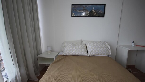 Une chambre à coucher comprenant un lit, une table de chevet, une commode, une fenêtre avec des rideaux et un tableau accroché au mur.
